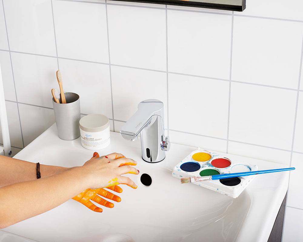 Kinderhände, die mit orangefarbenem Lack bedeckt sind, werden an einem Badezimmerbecken gewaschen, mit einer Farbpalette und einem Pinsel in der Nähe.