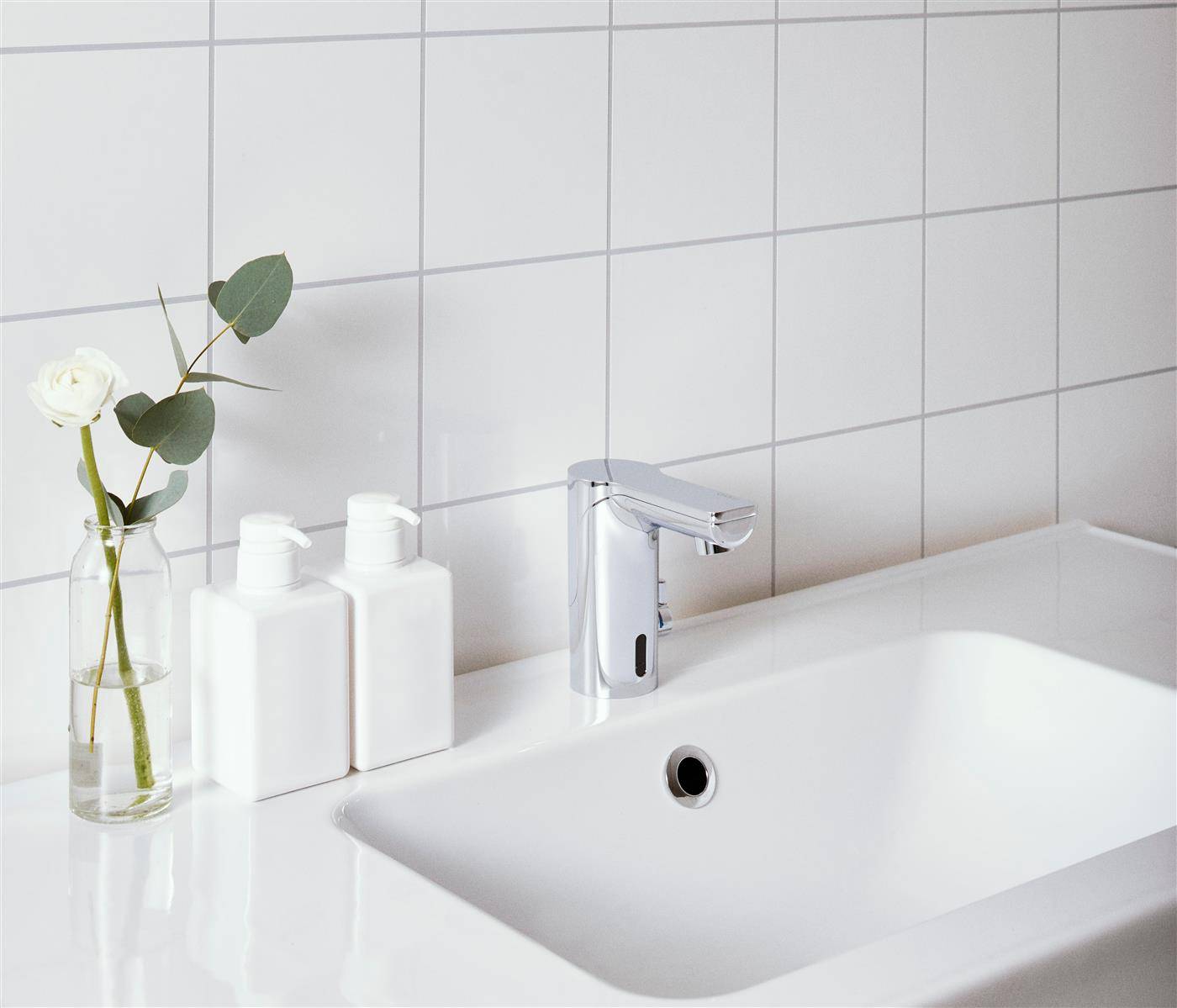 Modernes weißes Badezimmerbecken mit Chromarmatur, zwei weiße Flaschen und eine Vase mit einer weißen Rose und Grünpflanzen auf der Arbeitsplatte.