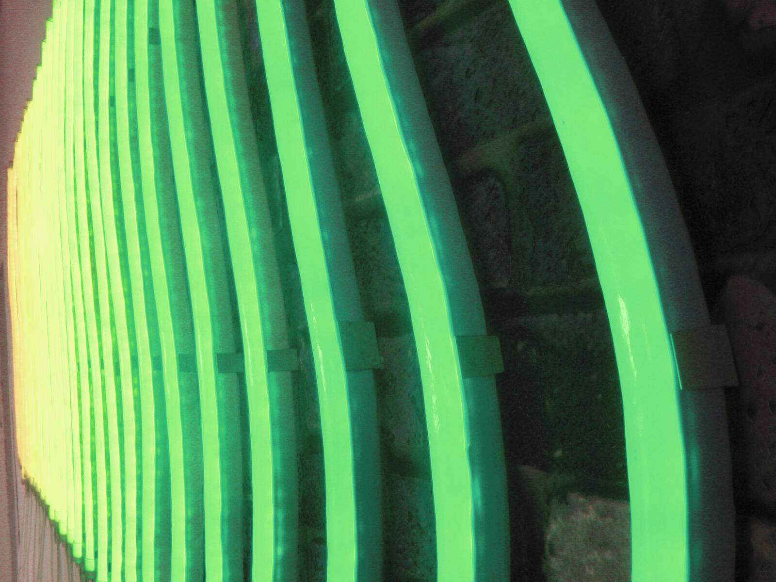 Grüne Neonlichter wölben sich gegen eine dunkle Backsteinmauer und erzeugen ein lebendiges, welliges Muster.