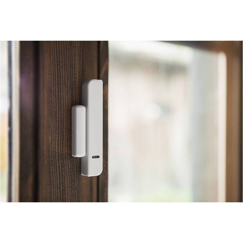 Weißer Tür- oder Fenstersensor auf einem dunklen Holzrahmen neben einer Glasscheibe montiert, wahrscheinlich zur Haussicherung oder Überwachung.
