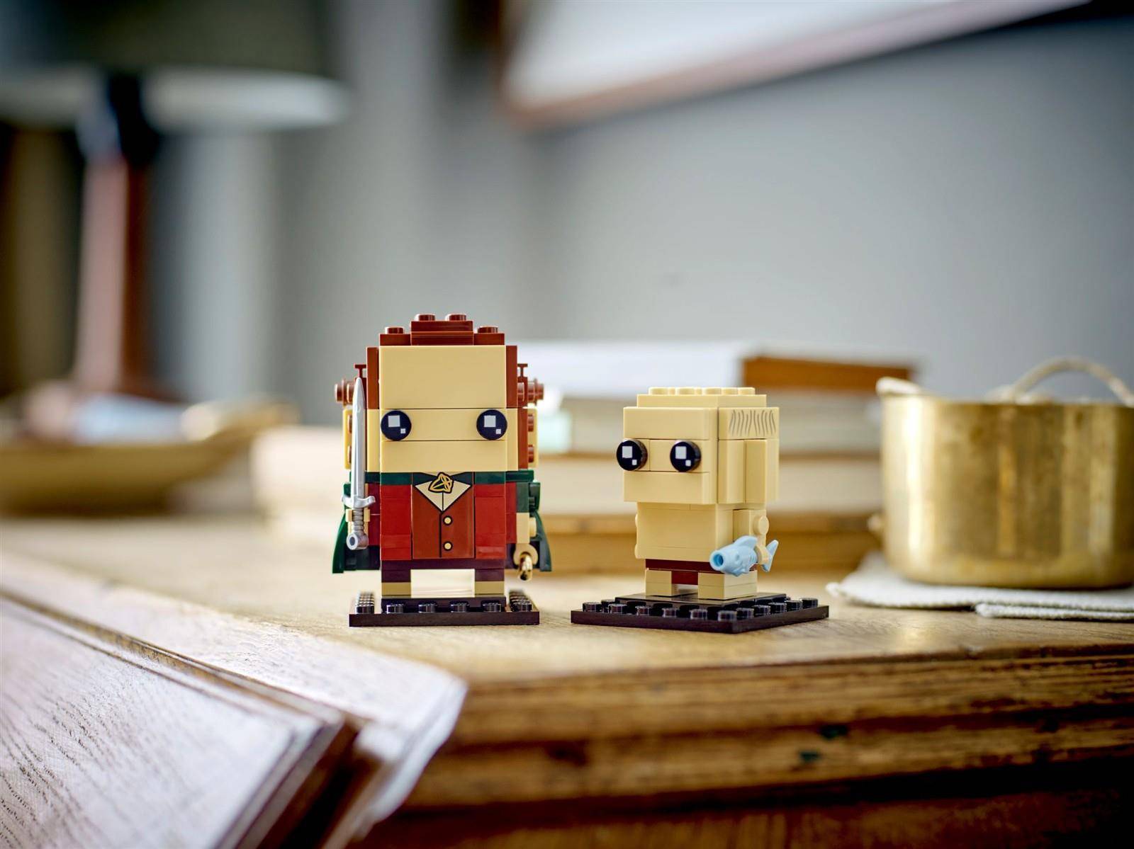 Zwei kleine LEGO-ähnliche Steinfiguren stehen auf einem Holztisch, mit einer unscharfen Tasse und einem Buch im Hintergrund.