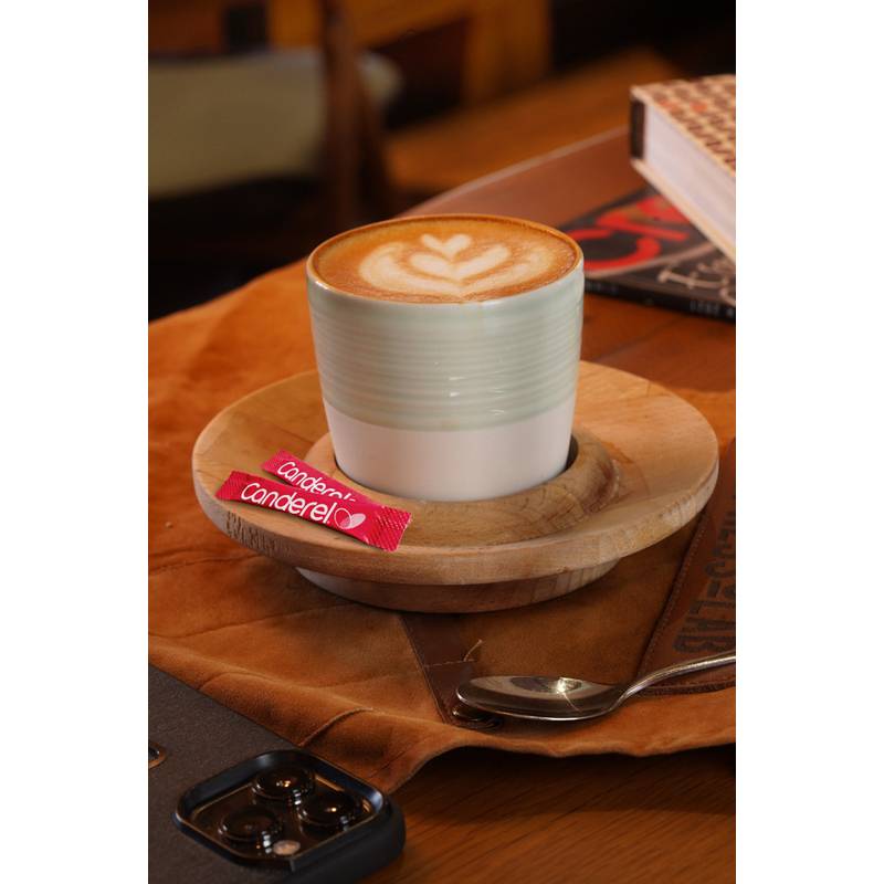 Eine Tasse Latte mit Latte-Kunst auf einer Holzuntertasse, neben einem roten Canderel-Zuckerersatzpäckchen und einem Löffel auf einem braunen Tisch.