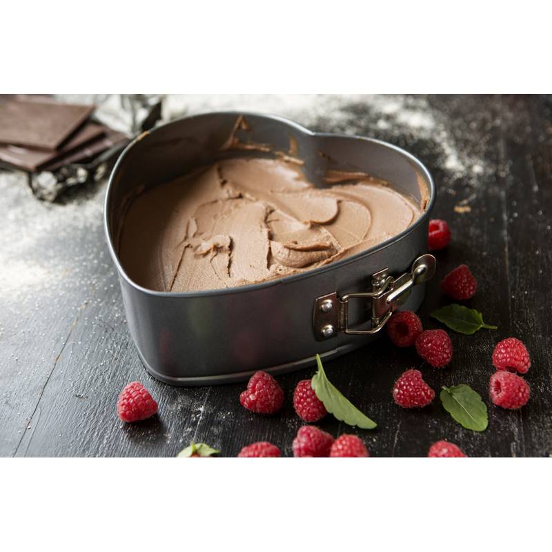Eine herzförmige Backform, gefüllt mit Schokoladenteig, umgeben von verstreuten Himbeeren und Minzblättern auf einer dunklen Holzoberfläche.