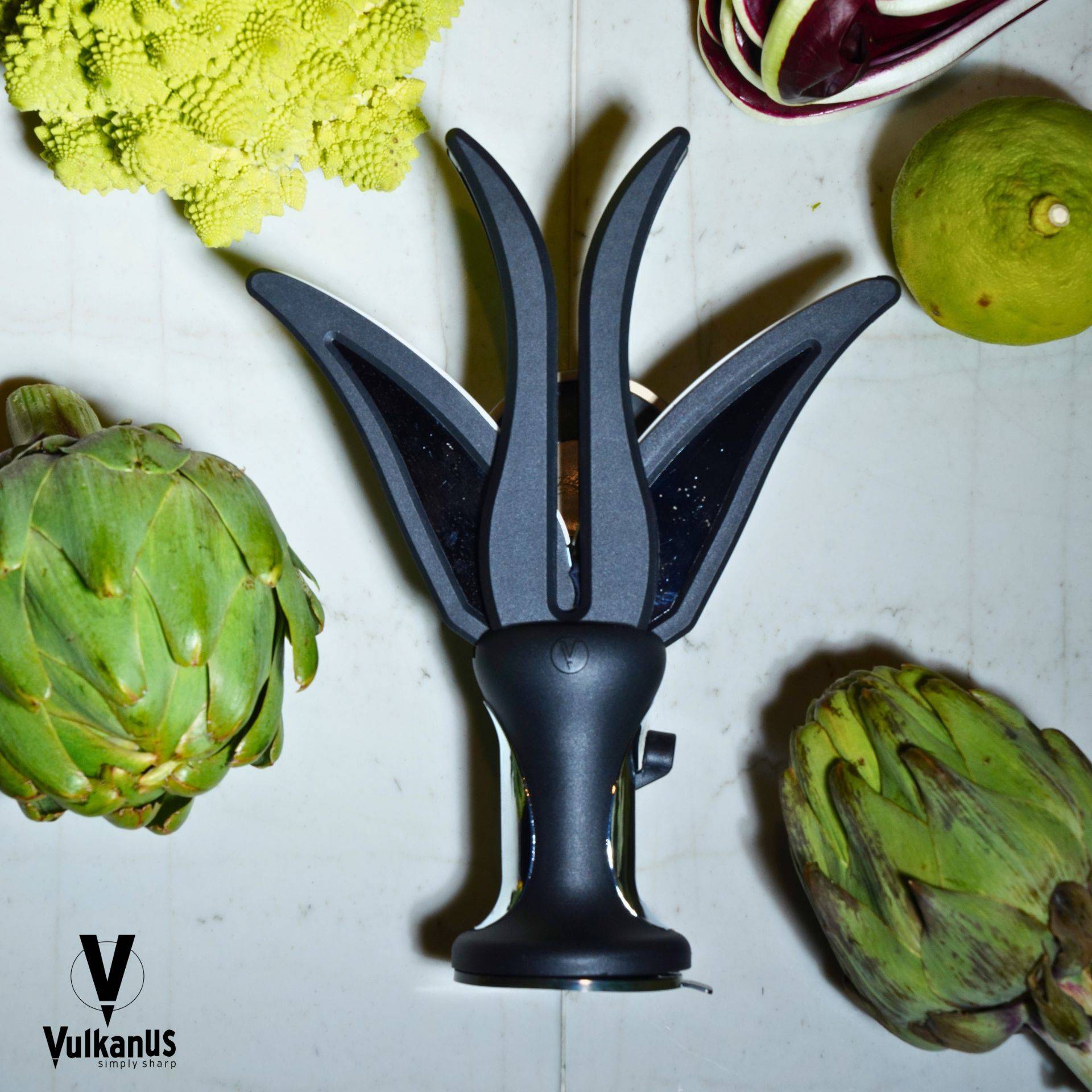 Ein Messerschärfer mit mehreren Klingen ist von verschiedenem Gemüse umgeben, einschließlich Artischocken, auf einer weißen Oberfläche.