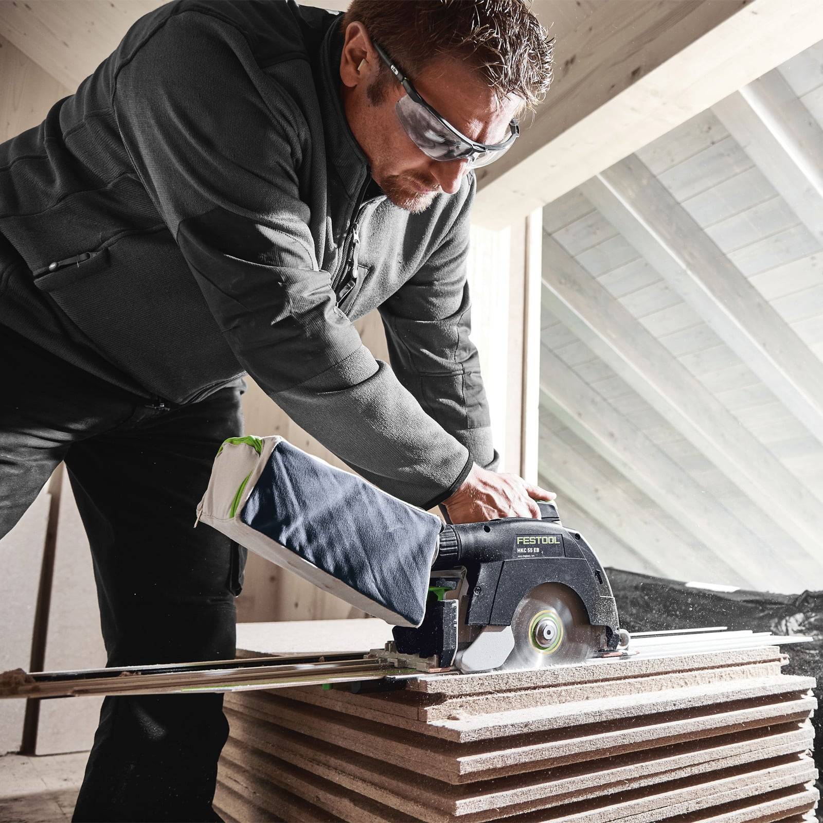 Eine Person mit Schutzbrille bedient eine Kreissäge und schneidet gestapelte Holzbretter in einer Werkstattumgebung.
