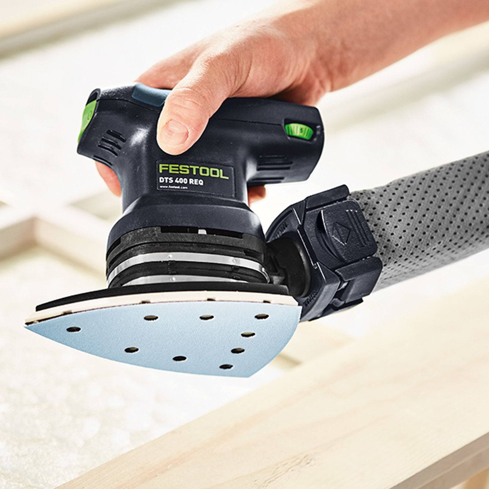 Eine Hand verwendet einen Festool DTS 400 REQ Schleifer und glättet eine Holzoberfläche. Das Gerät verfügt über ein kompaktes, ergonomisches Design mit einem Staubabsaugungsaufsatz.