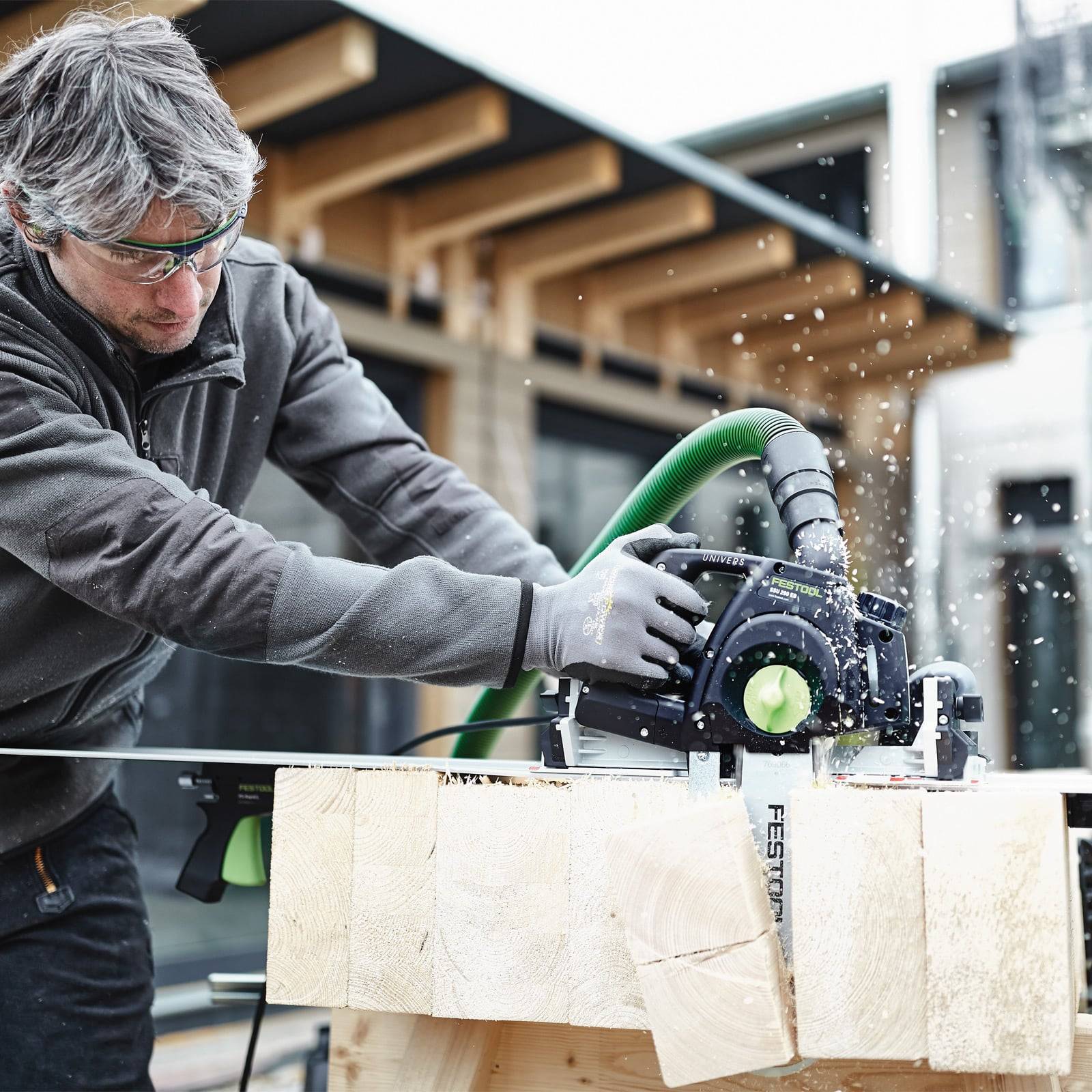 Eine Person mit Sicherheitsausrüstung sägt einen Holzbalken mit einer Kreissäge in einer Werkstattumgebung, umgeben von Holzkonstruktionen.