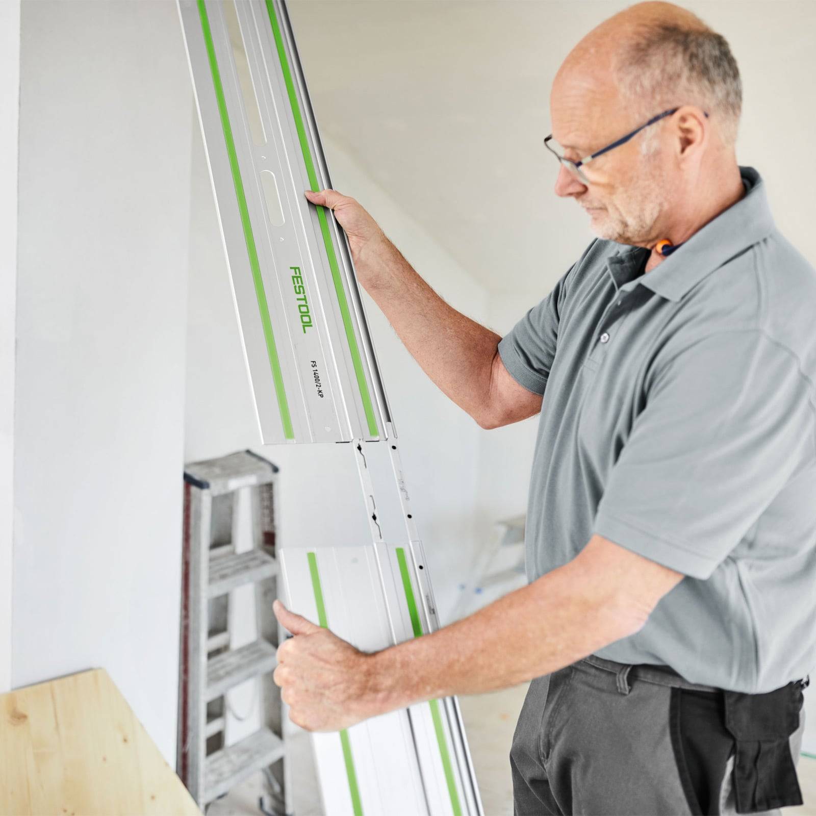 Eine Person mit Brille inspiziert ein langes Metallwerkzeug oder eine Führungsschiene mit 'Festool' Markierung und steht dabei in einem hell erleuchteten Raum.