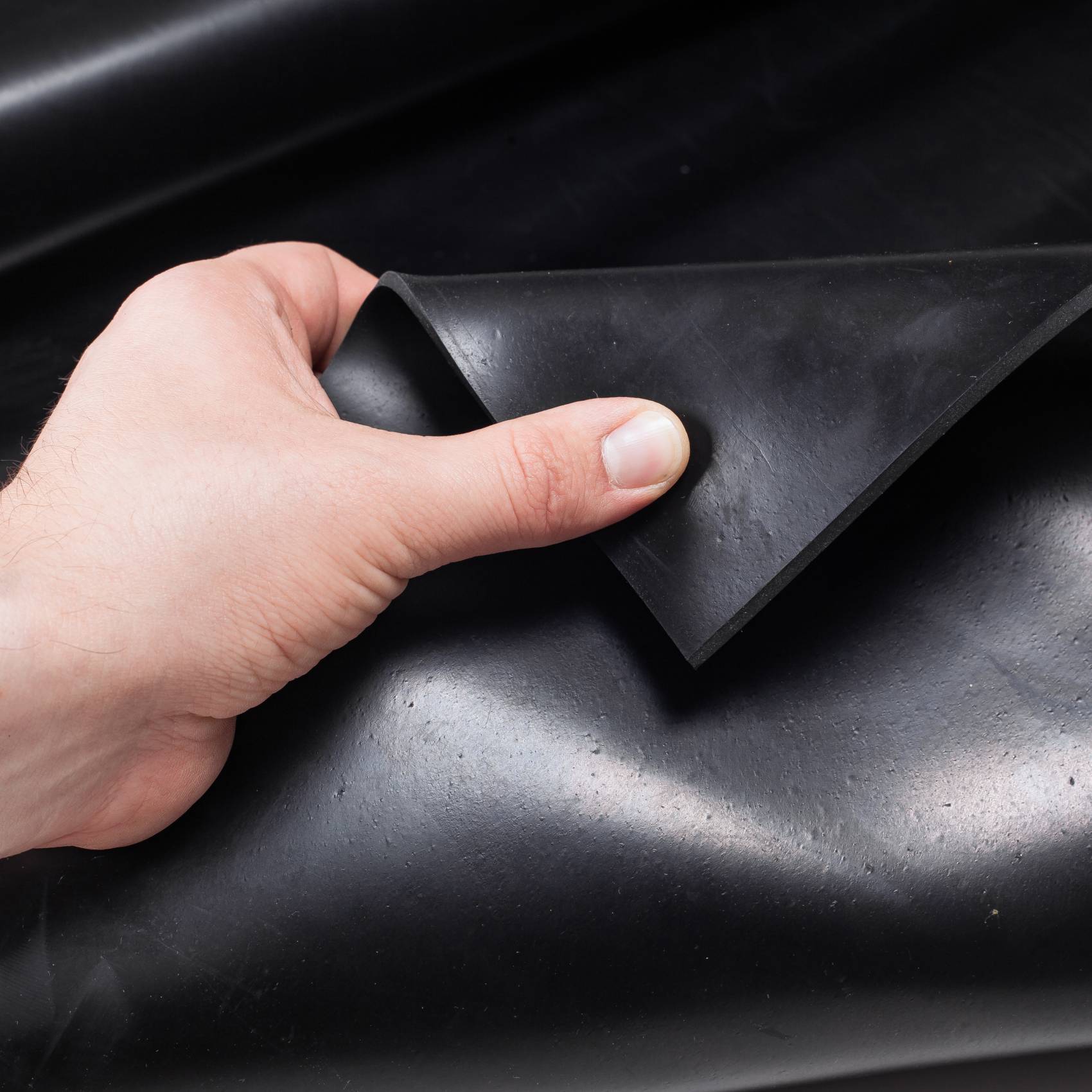 Hand hebt eine Ecke einer dicken schwarzen Gummimatte, wobei ihre flexible, strukturierte Oberfläche sichtbar wird.