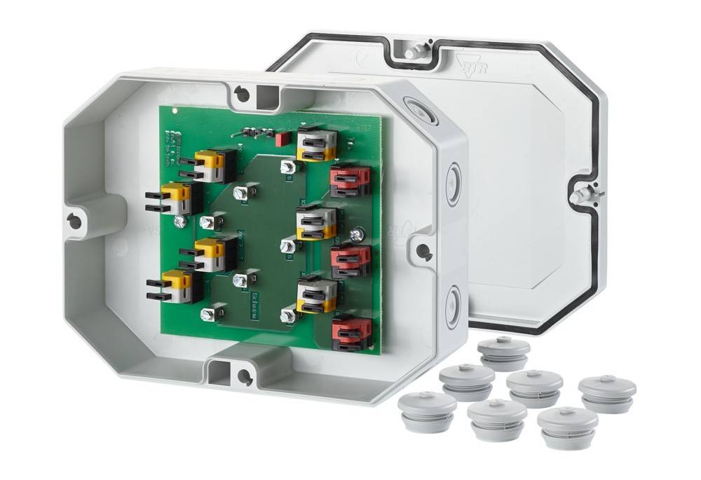 Sechseckige Anschlussbox mit grüner Leiterplatte im Inneren, mit mehreren beschrifteten Anschlüssen. Deckel der Box und mehrere Stecker separat dargestellt.