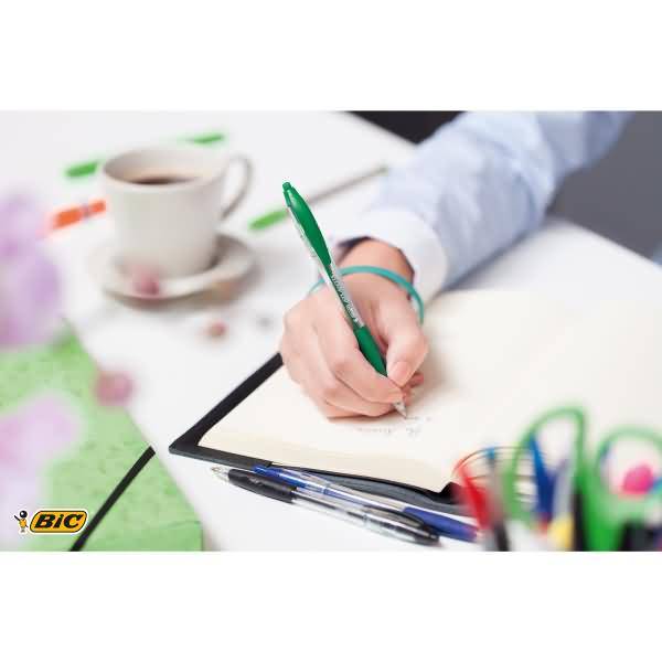 Nahaufnahme einer Person, die mit einem grünen Stift in einem Notizbuch an einem Schreibtisch schreibt, mit einer Kaffeetasse und Stiften in der Nähe.
