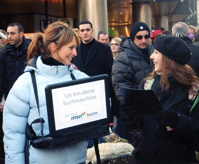 Zwei Frauen, die auf einer belebten Straße stehen und miteinander lächeln. Eine von ihnen hält einen Laptop, auf dessen Bildschirm der Text 