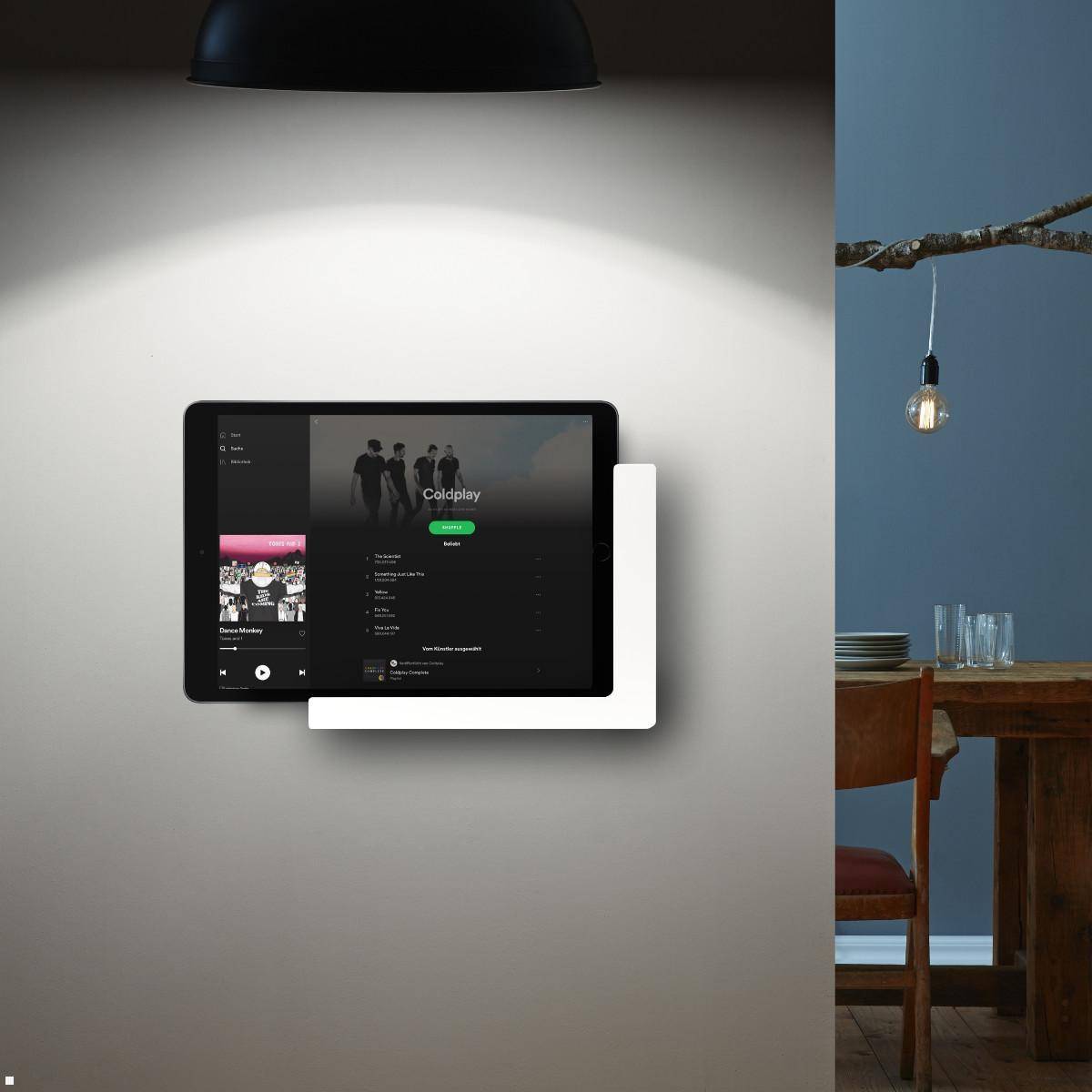 Eine an der Wand montierte Tablette zeigt eine Musik-Streaming-App mit einer Playlist, während im Hintergrund ein Essbereich zu sehen ist.