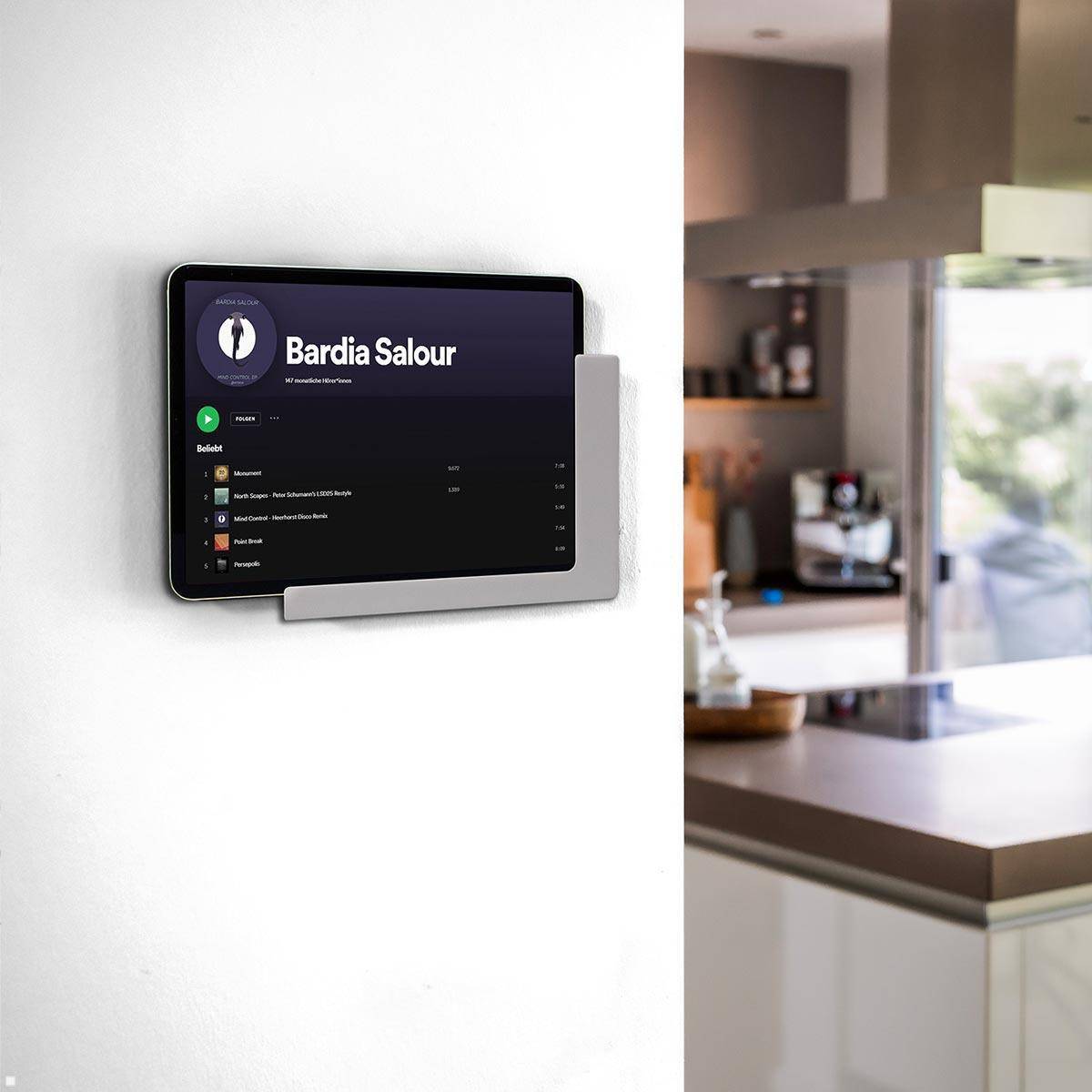 Anzeige zur Smart-Home-Steuerung an der Wand, die eine Musikliste von Bardia Salour zeigt. Moderne Küche im Hintergrund sichtbar.