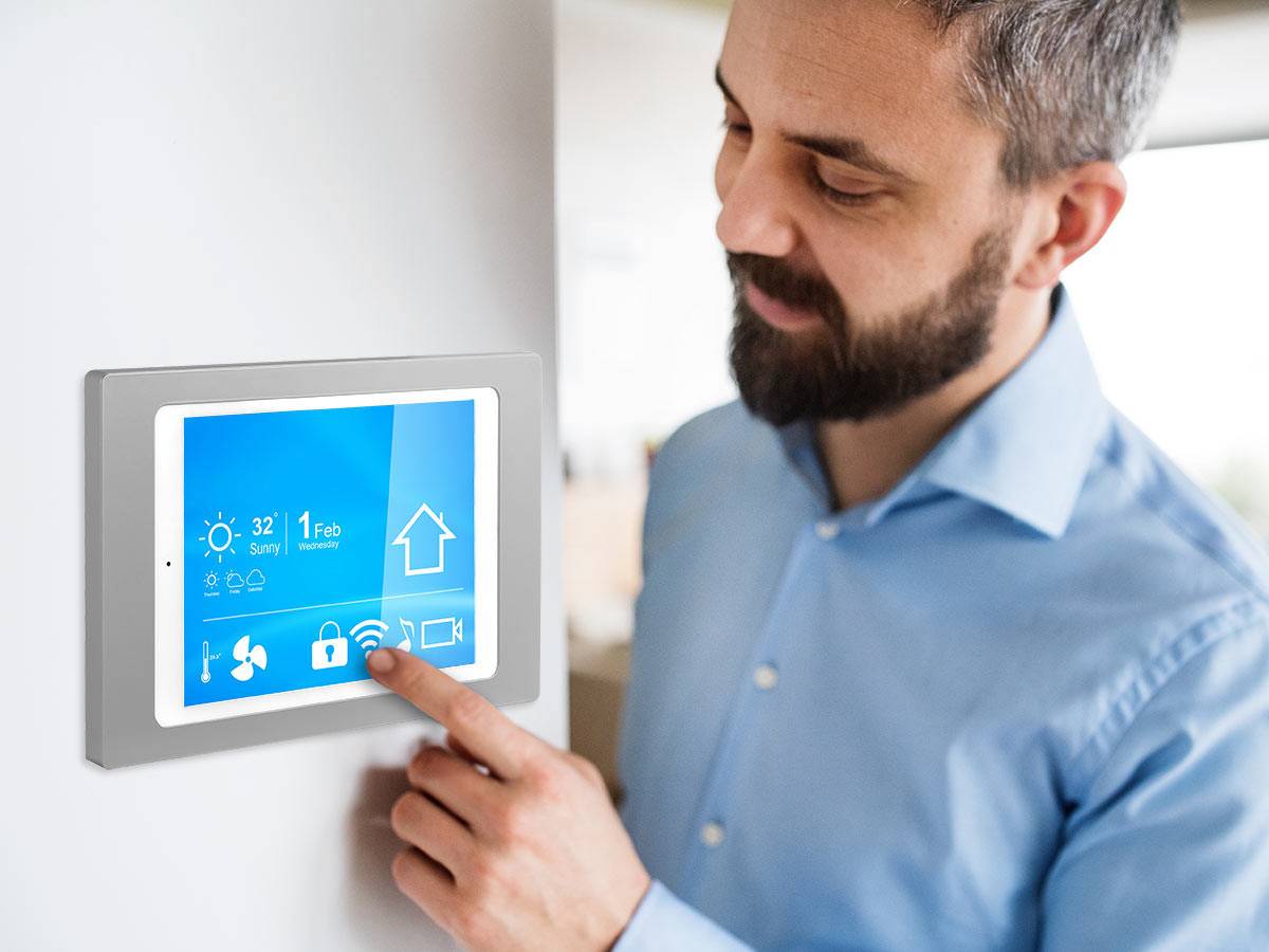 Ein Mann interagiert mit einem intelligenten Heimsteuerungspanel, das Wetter-, Sicherheits- und Konnektivitäts-Icons zeigt und damit eine moderne Technologie-Umgebung andeutet.