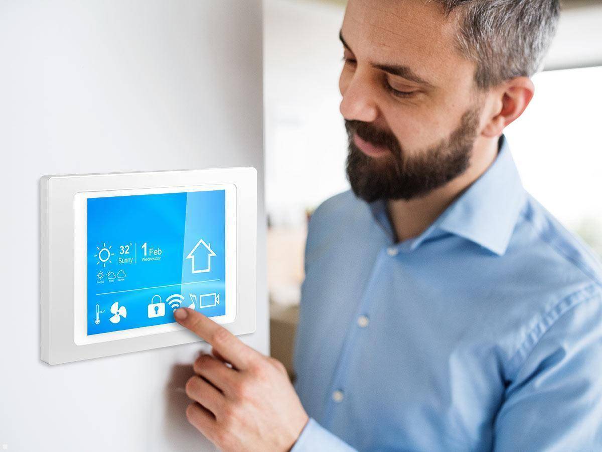 Eine Person interagiert mit einem Hausautomationspanel, das Wetter, Temperatur, Kalenderdatum und verschiedene Steuerungssymbole anzeigt.