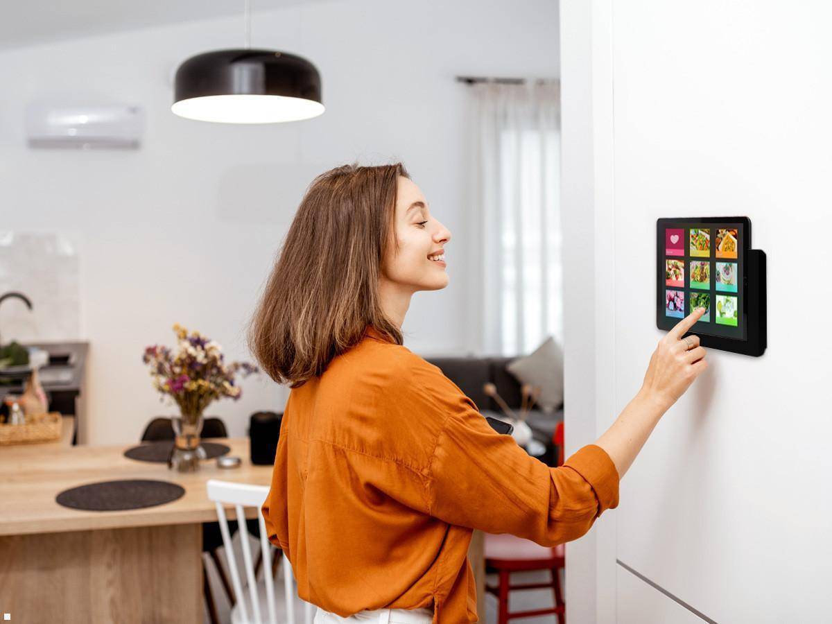 Eine Frau in einem orangefarbenen Shirt interagiert mit einer digitalen Fotoausstellung an der Wand in ihrem Zuhause. Ein Esstisch und Stühle sind im Hintergrund sichtbar.