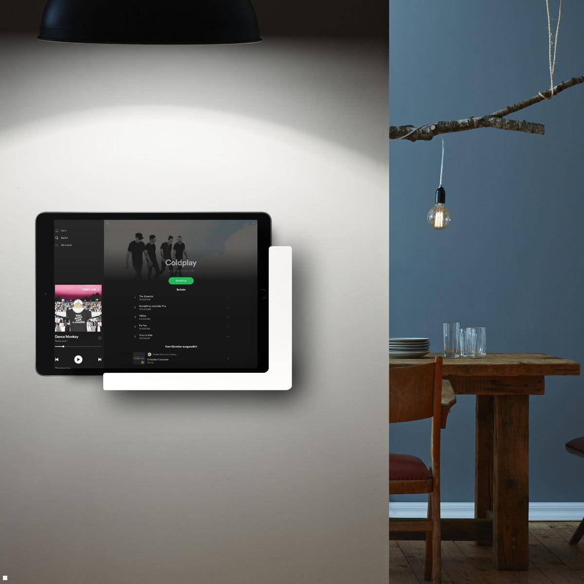 Eine auf einer weißen Wand befestigte Tablette zeigt eine Musik-App mit einer Playlist. In der Nähe sind ein Holztisch und Stühle teilweise zu sehen.