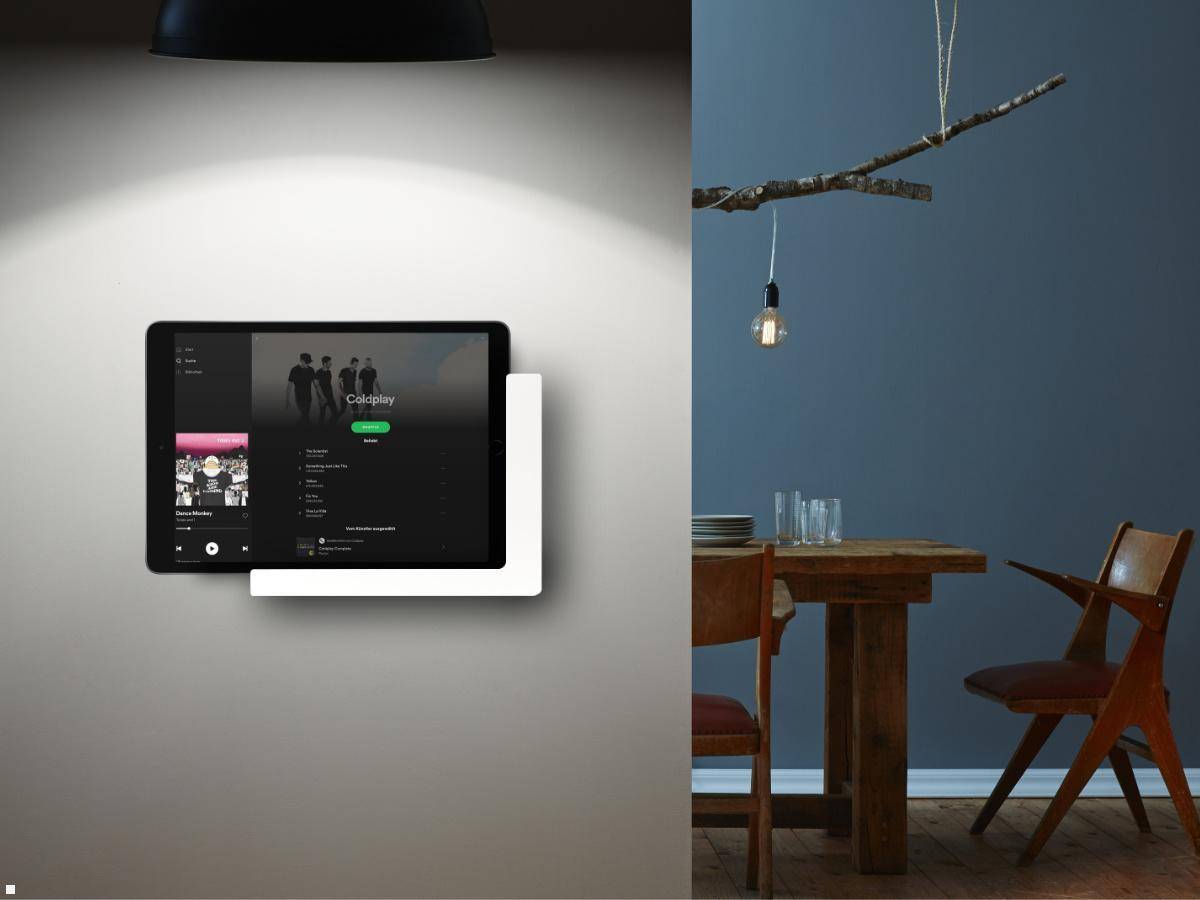 Eine Tablette mit einer Musik-App ist unter einer schwarzen Lampe an der Wand befestigt; rechts davon befindet sich ein Esstisch mit Stühlen, vor einer blauen Wand.