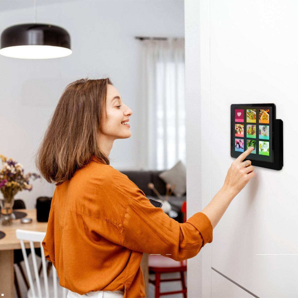 Eine Frau in einem orangefarbenen Shirt interagiert mit einer wandmontierten digitalen Anzeige, die farbenfrohe Bilder in einer Wohnumgebung zeigt.