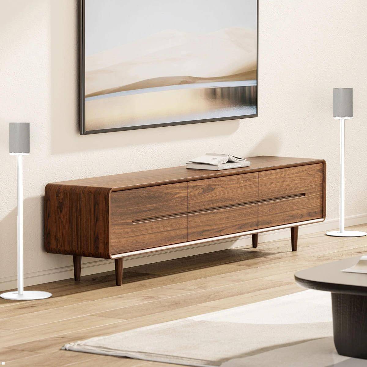 Ein Holzsideboard mit eleganten Linien steht an einer beigen Wand, auf der Oberseite mit Büchern bestückt. Zwei weiße Lautsprecher rahmen ein minimalistisches Landschaftsgemälde ein.