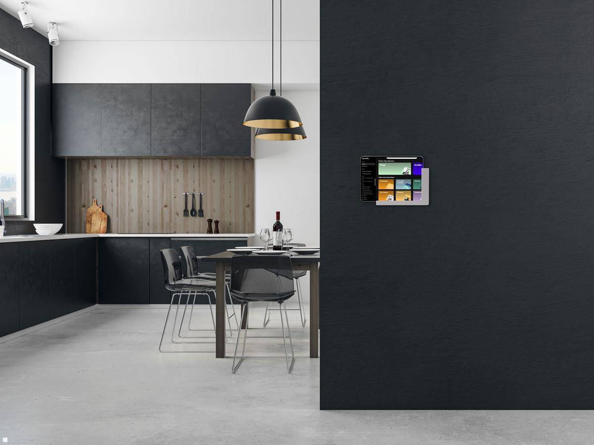 Moderne Küche mit dunklen Küchenschränken und einem Esstisch. Ein schlankes Tabletgerät ist an der angrenzenden grauen Wand montiert und zeigt verschiedene Apps an.