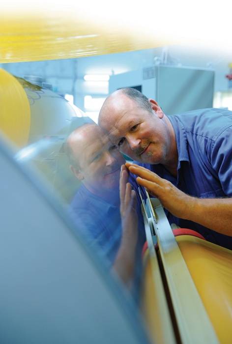 Ein Mann in einem blauen Hemd untersucht aufmerksam und konzentriert eine reflektierende Oberfläche in einer Fertigungsumgebung und zeigt dabei Präzision und Sorgfalt.