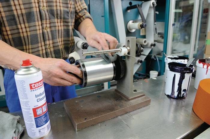 Eine Person, die einen Metallwalzenkörper mit einem Tuch in einer industriellen Umgebung reinigt. Ein Behälter mit der Aufschrift „Industriereiniger