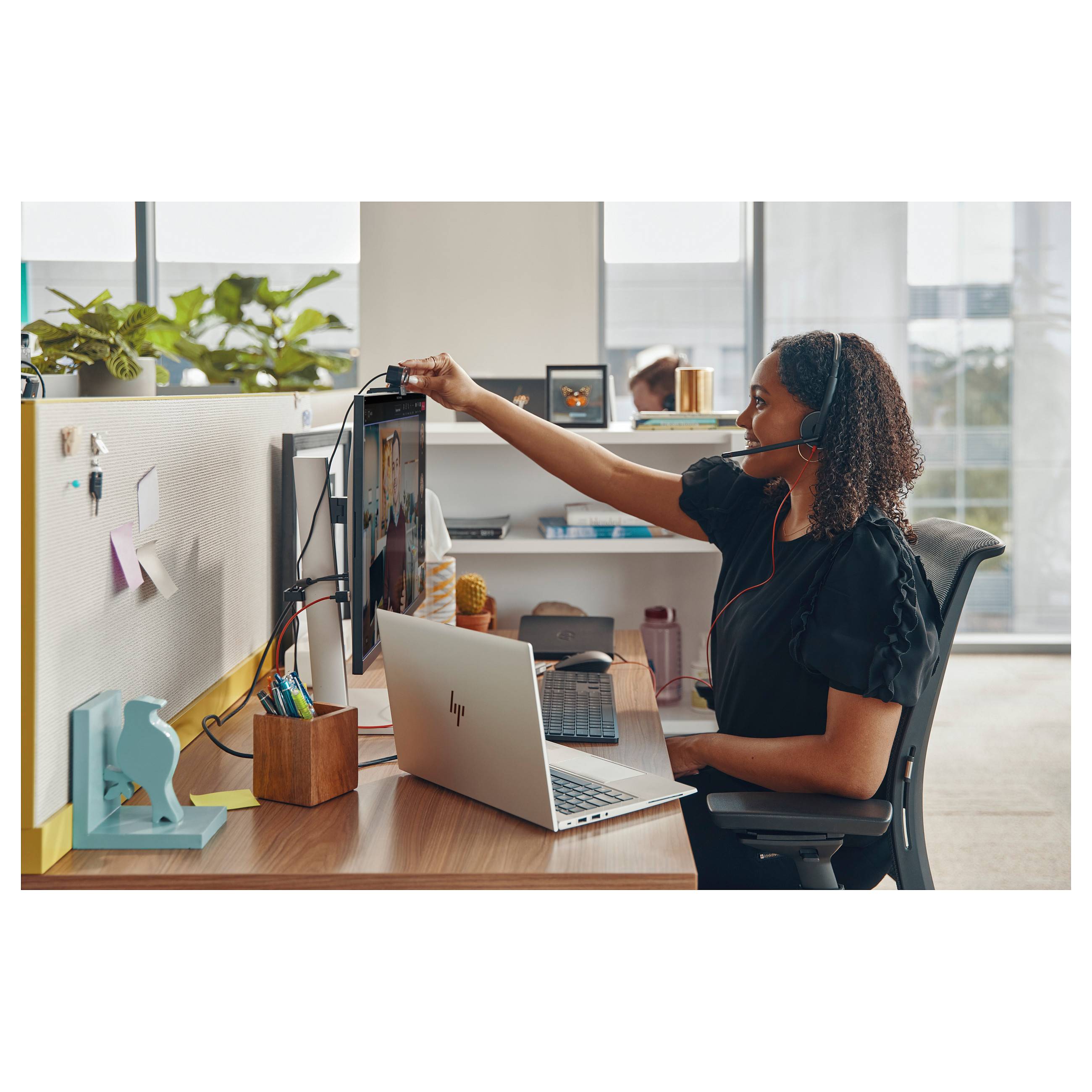 Eine Person mit Kopfhörer arbeitet an einem Schreibtisch mit einem Laptop und einem großen Monitor. Sie stellt die Webcam ein, während sie in einer Büroumgebung sitzt.