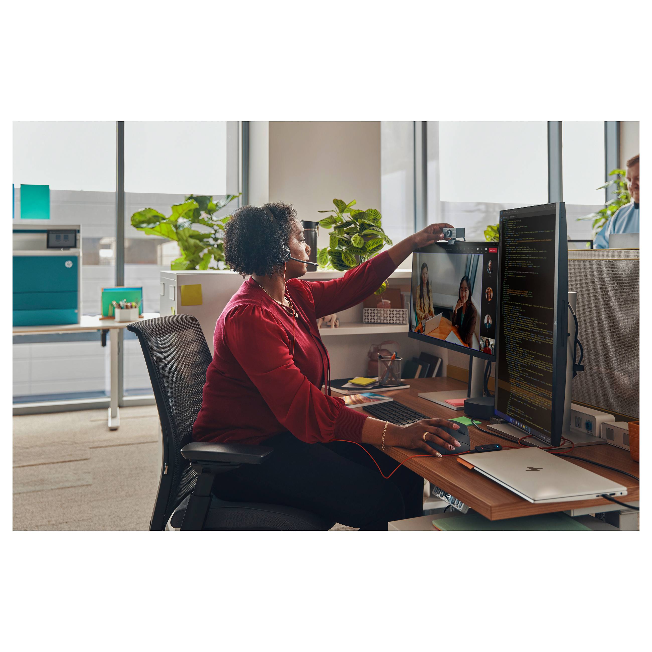 Eine Person im Büro mit einem Kopfhörer, die sich an einem Videoanruf auf einer Dual-Monitor-Einrichtung beteiligt, mit sichtbaren Pflanzen und Büromaterialien.