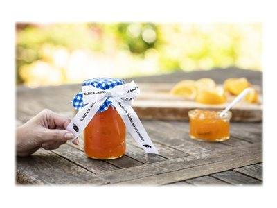 Eine Person hält ein Glas Orangenmarmelade, das mit einem Band verschlossen ist, auf einem Holztisch. Im Hintergrund befindet sich eine kleine Schale mit Marmelade und geschnittenen Orangen.