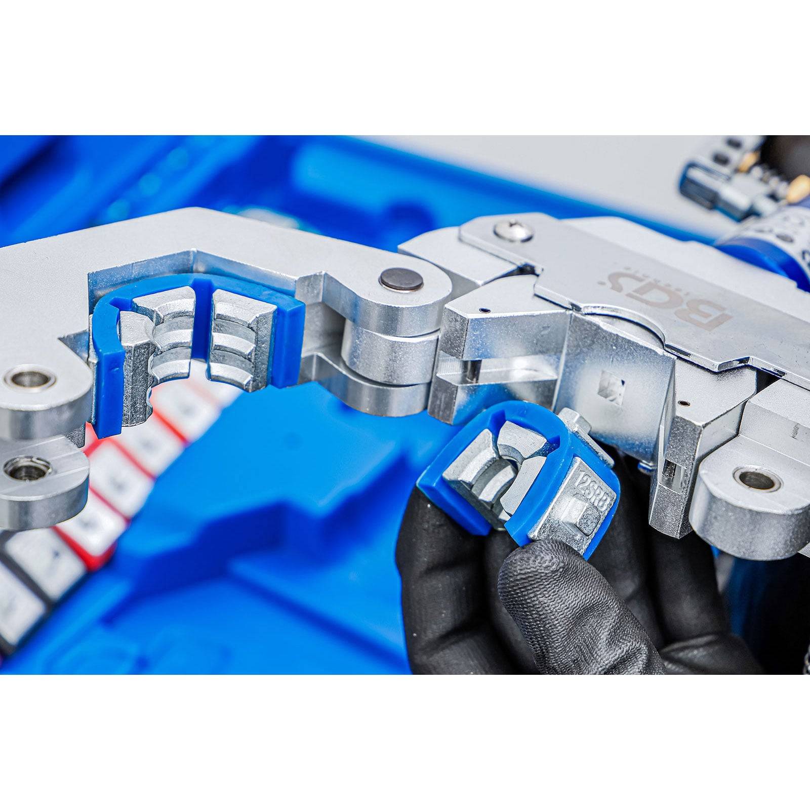 Eine behandschuhte Hand hält ein kleines Metallwerkzeugteil mit blauem Gehäuse, das neben einem größeren mechanischen Gerät auf blauem Hintergrund gezeigt wird.