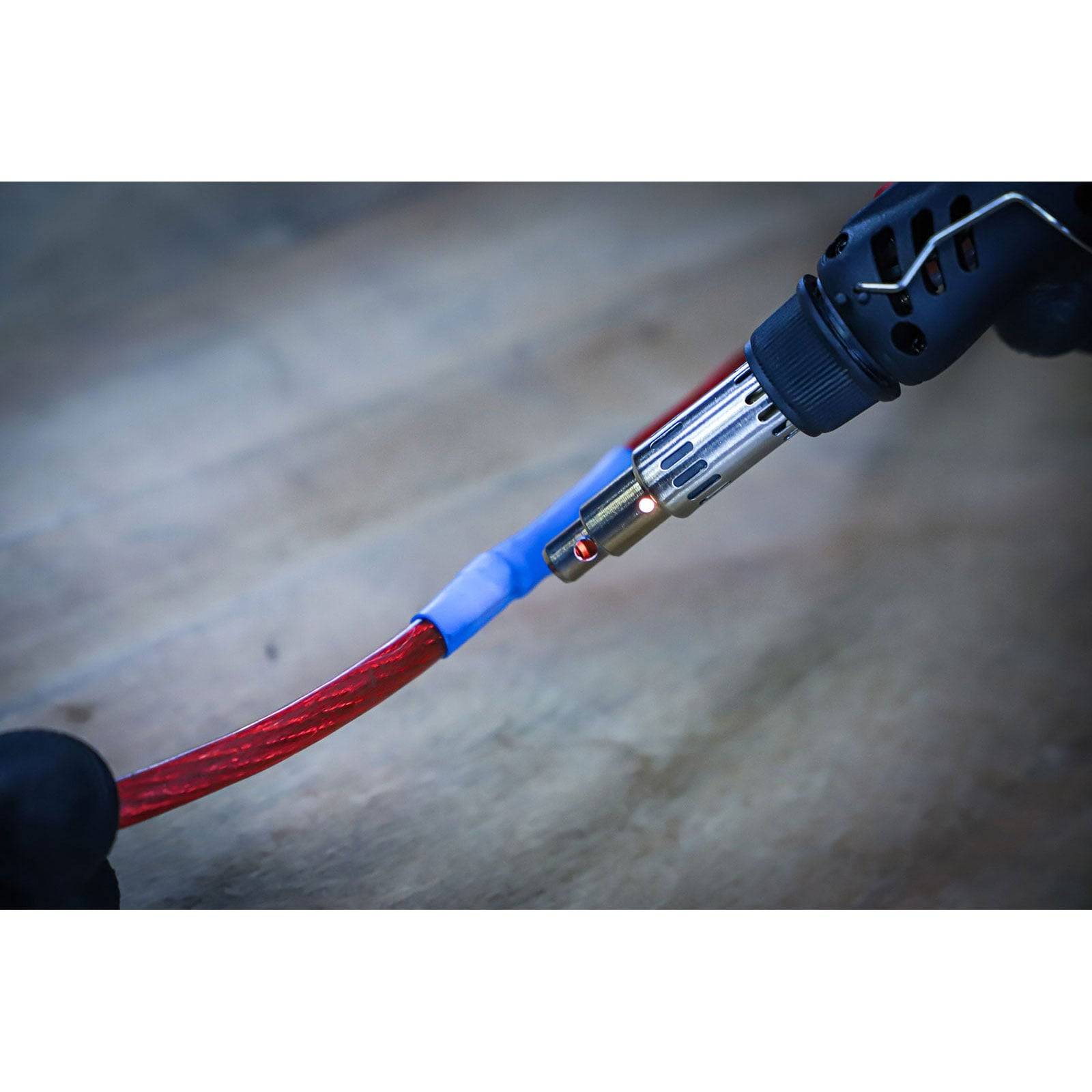 Eine Hand hält einen Heißluftfön und schrumpft blaues Schrumpfschlauchrohr um ein rotes Kabel auf einer Holzoberfläche und demonstriert dabei die Kabelisolierung.