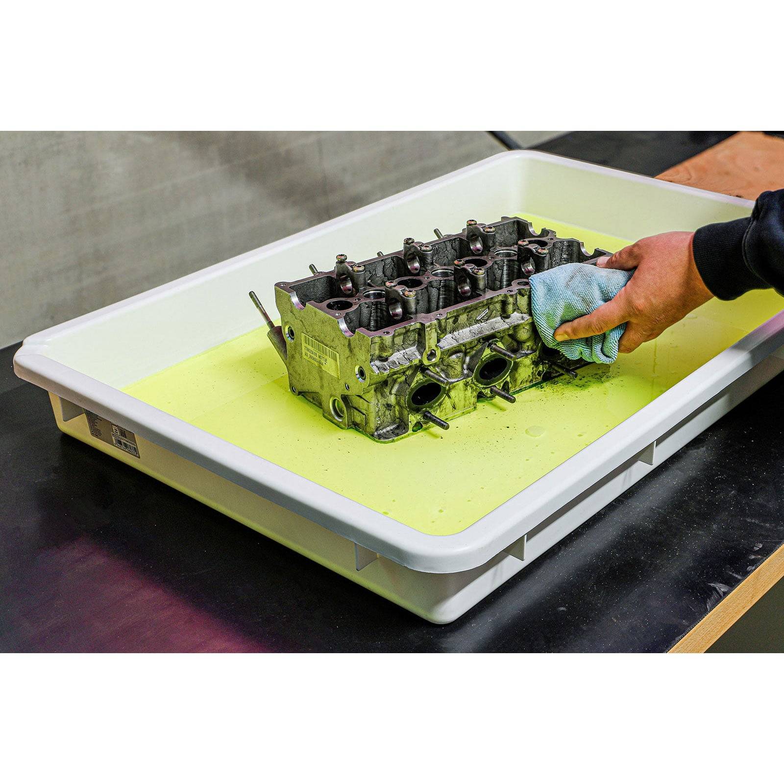 Eine Person reinigt ein Metallteil eines Motors, das in einem Tablett mit gelber Auskleidung liegt und dabei ein blaues Tuch verwendet. Die Umgebung ist ein industrieller Arbeitsbereich.