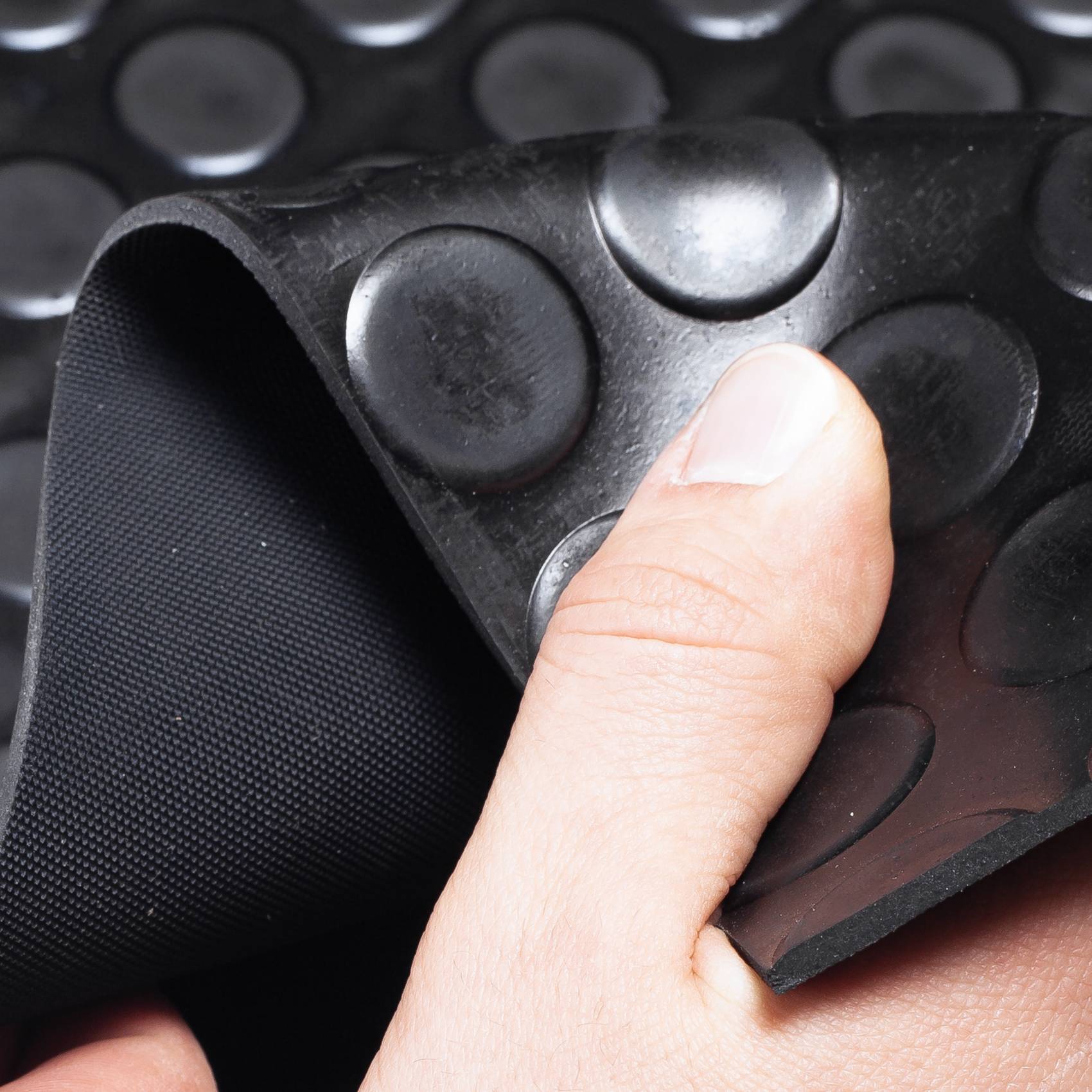 Hand, die eine Ecke einer schwarzen Gummimatte mit runden erhabenen Noppen zurückzieht, um die strukturierte Unterseite zu zeigen.