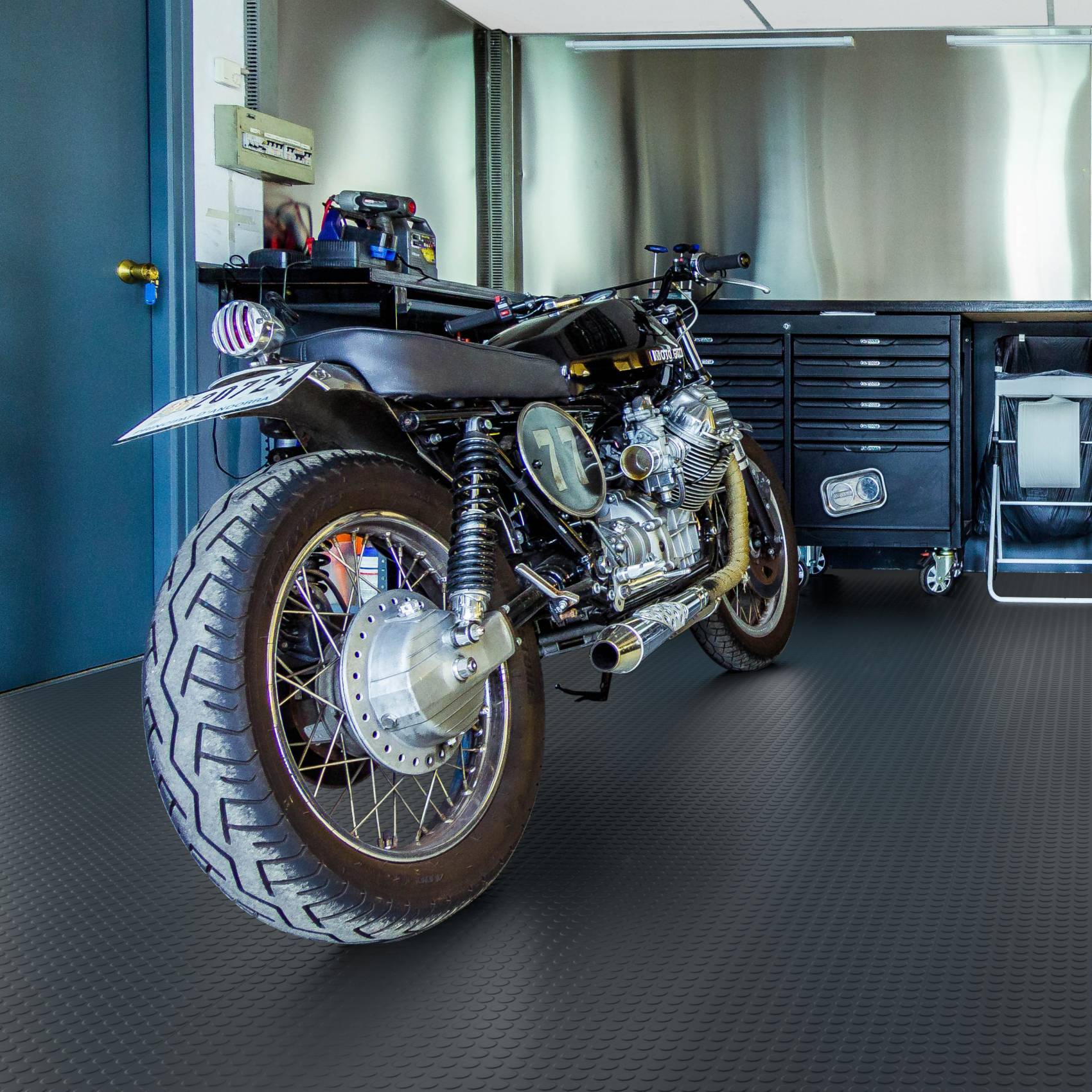 Schwarzes Custom-Motorrad, das in einer Garage oder Werkstatt geparkt ist, aus der hinteren linken Perspektive fotografiert, mit Werkzeugschränken im Hintergrund.