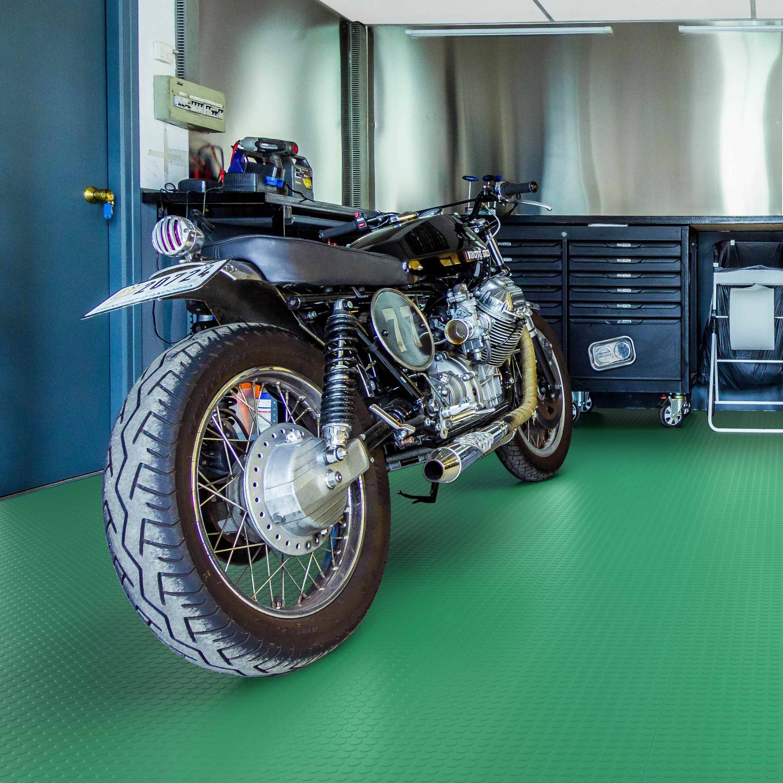 Ein maßgefertigtes schwarzes Motorrad in einer Garage, von hinten-links gezeigt, auf einem grünen Boden neben Werkzeugschränken.