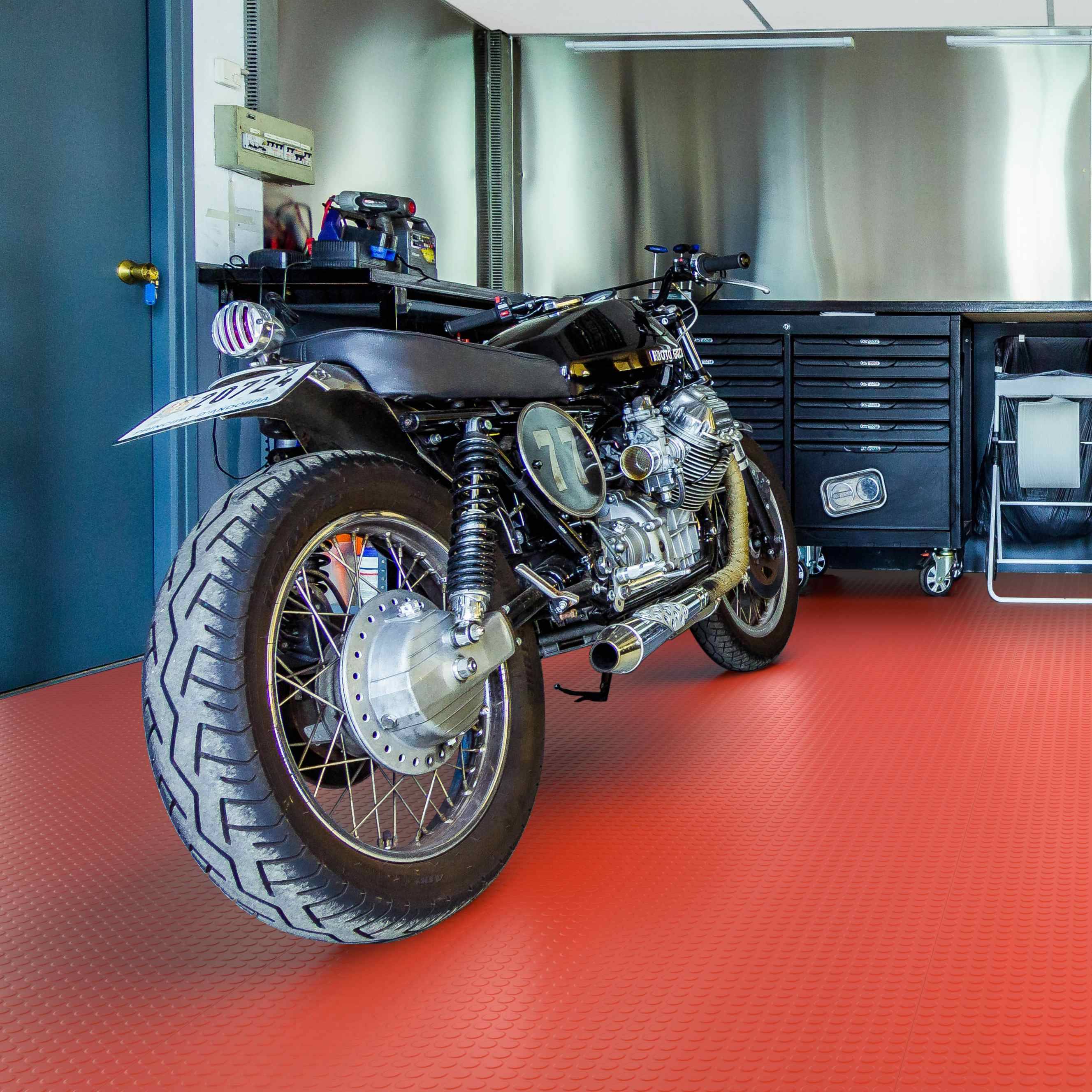 Ein benutzerdefiniertes schwarzes Motorrad parkt in einer Garage auf einem roten Boden, mit Werkzeugschränken und einer Werkbank im Hintergrund.