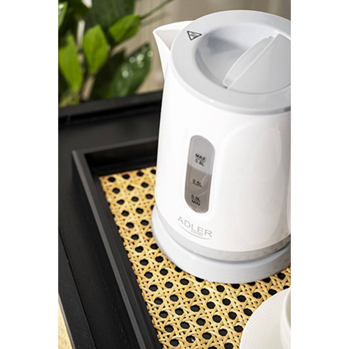 Elektrischer Wasserkocher auf einem gewebten Tablett neben einer Pflanze. Der Wasserkocher zeigt die Wassermenge in Litern angezeigt.