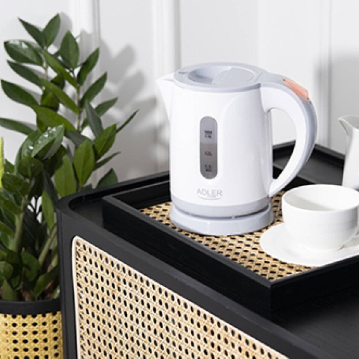 Elektrischer Wasserkocher auf einem Tablett neben einer Teetasse, mit Pflanzen im Hintergrund.