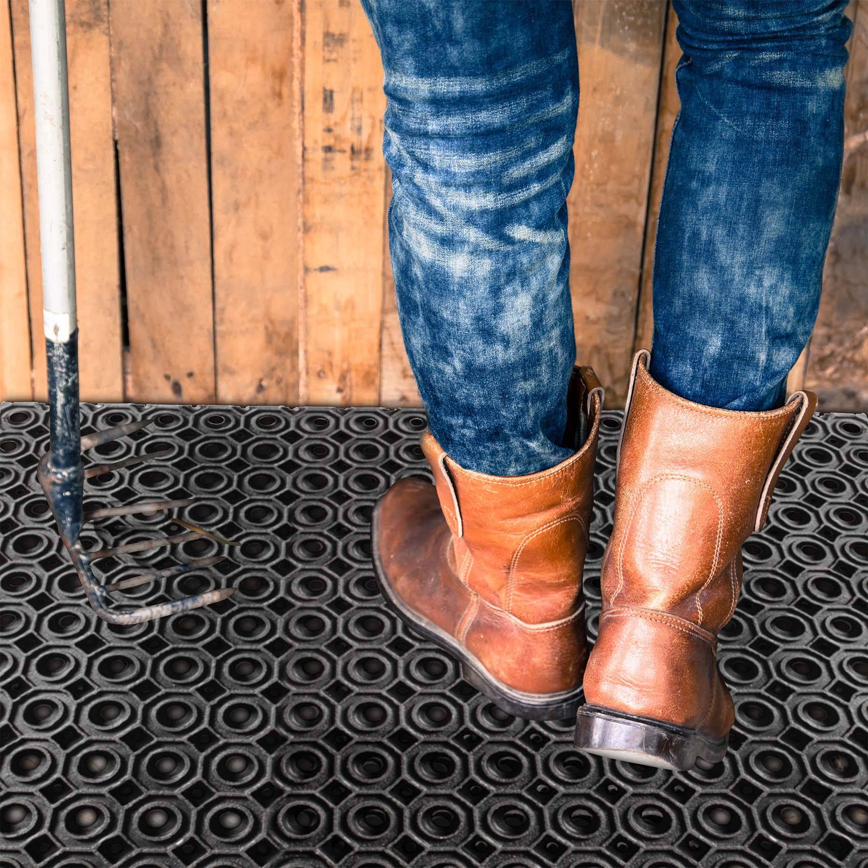 Eine Person steht auf einer schwarzen Gummimatte und trägt verblichene Jeans und braune Lederstiefel vor einer Holzwand.