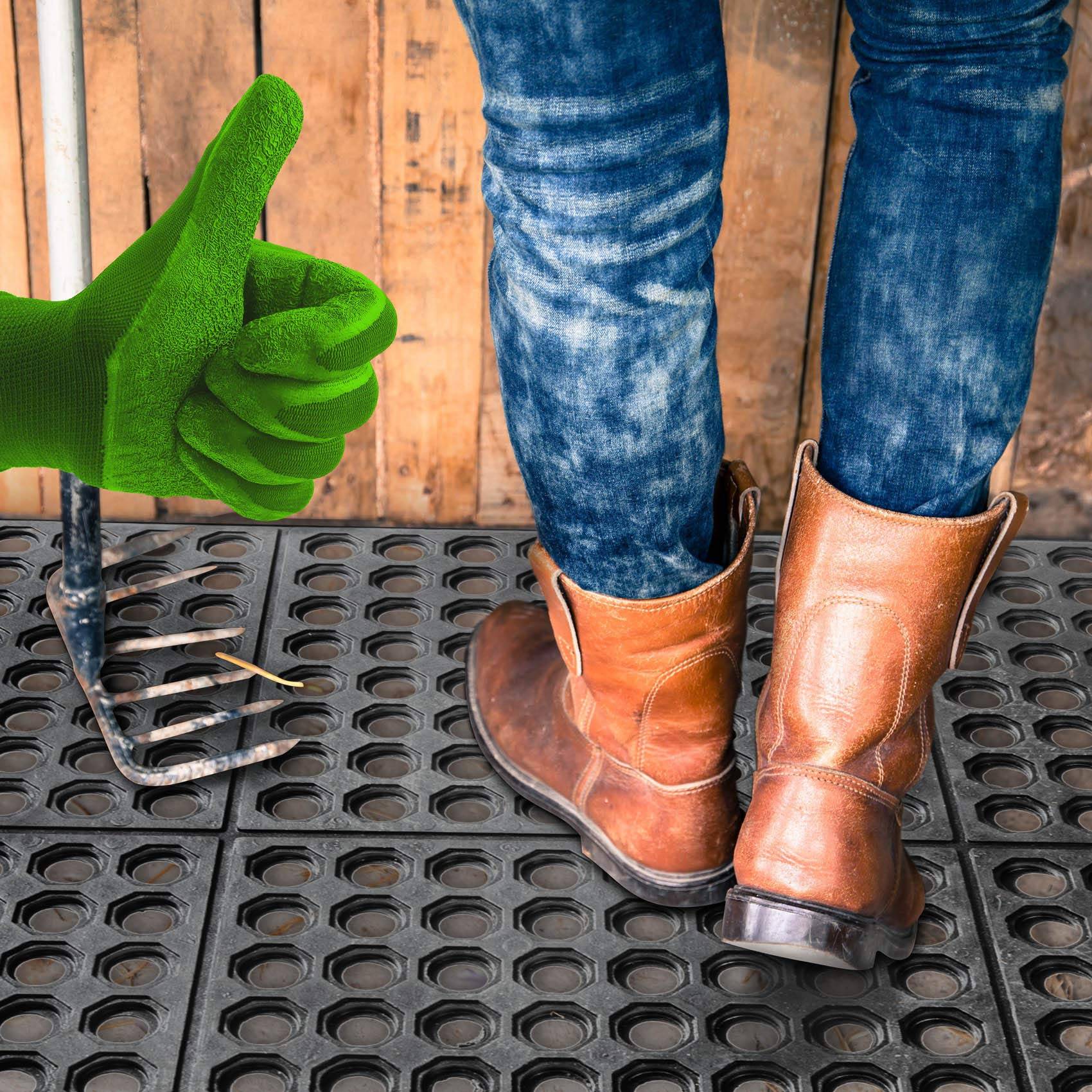 Grün behandschuhte Hand gibt einen Daumen hoch neben einer Gartenheugabel, neben einer Person in Jeans und braunen Arbeitstiefeln, die auf schwarzen Gummimatten steht.