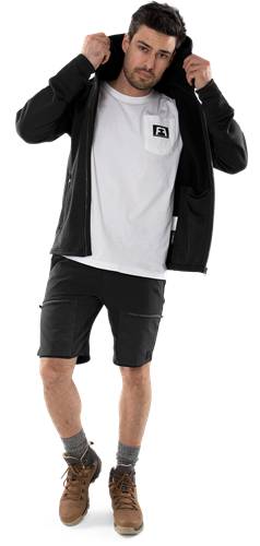 Eine Person trägt eine schwarze Jacke über einem weißen Hemd und schwarzen Shorts und steht vor einem einfarbigen schwarzen Hintergrund.