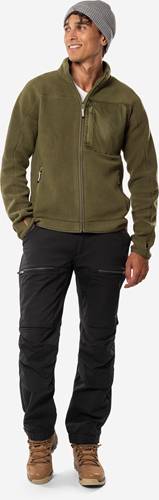 Eine Person in einer grünen Jacke mit Reißverschluss und schwarzer Hose, die die Hände in den Taschen hat, steht vor einem neutralen Hintergrund.