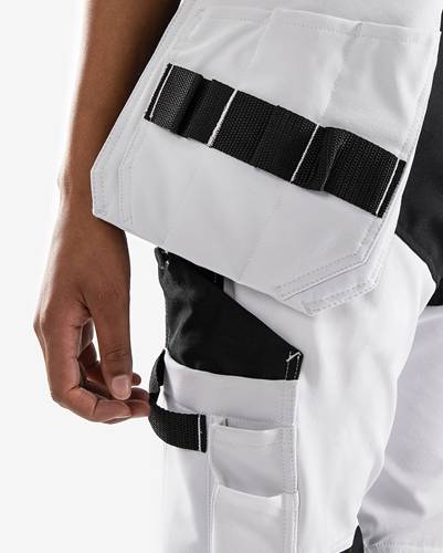 Eine Person in einer weißen Uniform mit schwarzen Trägern steht vor einem einfarbigen Hintergrund und zeigt eine Nahaufnahme ihrer linken Hand und eines Teils der Uniform.