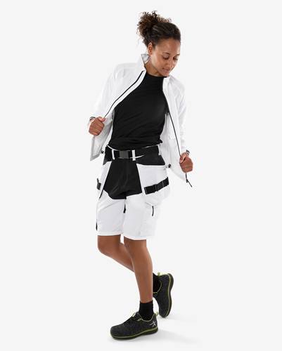 Eine Person in Sportkleidung posiert selbstbewusst und trägt ein schwarzes Oberteil, weiße Shorts mit Taschen und eine weiße Jacke. Sie hat lockiges Haar.