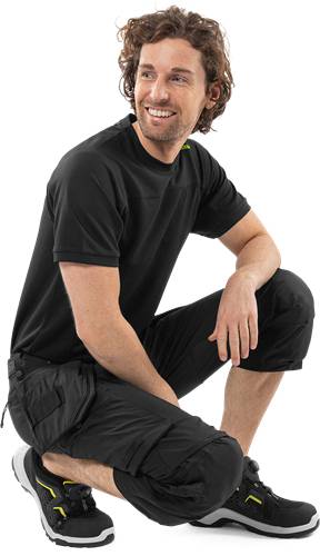 Eine Person in einem schwarzen Shirt und einer schwarzen Hose kniet mit verschränkten Armen und wirkt nachdenklich vor einem schlichten Hintergrund.