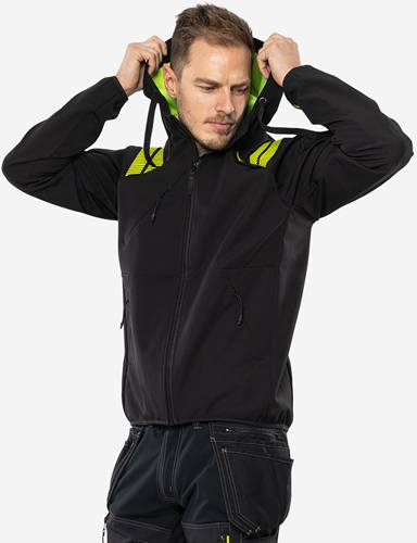 Eine Person trägt eine schwarze und neongrüne Jacke und richtet die Kapuze, vor einem schlichten Hintergrund.