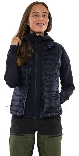 Eine Person trägt eine marineblau gesteppte Weste über einer schwarzen Jacke mit Reißverschluss, während sie mit der linken Hand ihr Haar berührt.