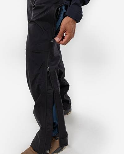 Eine Person schließt den Reißverschluss an der Seite einer schwarzen wasserdichten Hose über einer blauen Hose, was die Funktionalität für einfachen Zugang und Schichtung in Wanderausrüstung demonstriert.